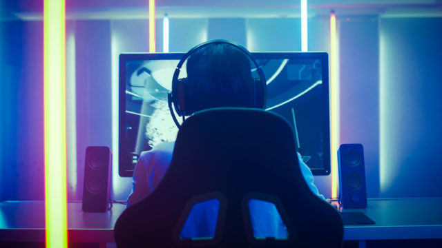 Back View Shot of the Professional Gamer Playing in First-Person Shooter Online Video Game on His Personal Computer. Room Lit by Neon Lights in Retro Arcade Style. Online Cyber e-Sport Internet Championship.